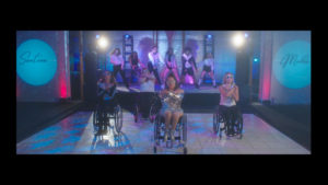 A charismatic woman who uses a wheelchair is center stage on the dance floor of a night club. She wears a sequined outfit and performs choreographed dance moves with three other women who use wheelchairs. Behind them, on a raised platform six other dancers of various races and genders dance. On either side of the dance floor, we see the woman’s named spelled out in lights: Santina Muha.