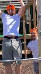 A bearded Caucasian male appears to be doing a pull up on a ladder rung of monkey bars while off to the side a male Caucasian Little Person is looking forward and smiling. Both are wearing the same outfit: grey shorts, white t-shirt, and an orange baseball cap.