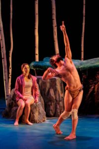 On a theater stage arranged with large boulders and birch trees a Caucasian woman with dark hair is sitting on a rock watching a Caucasian bearded man with faun horns dance with one arm reaching to the air.