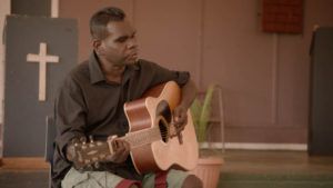 Geoffrey Gurrumul Yunupingu, an indigenous artist blind since birth, sits on a wooden chest holding his guitar. He has a dark grey photography scrim hanging behind him. The title of the film: GURRUMUL is at the bottom of the poster.
