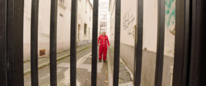 Through the thick black bars of a security gate, an older bald Caucasian man wearing a red track suit is using a cane as he walks down an empty graffiti marked alleyway.