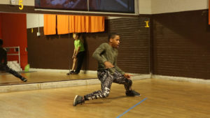 A young African-American boy is in the midst of a dance move at a dance studio. The mirror wall behind him reflects a young Caucasian boy with one of his pants' leg drawn up and is standing against a wall watching the young African-American boy dance.
