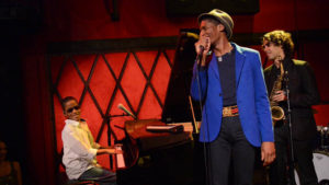 A young African-American boy wearing glasses is playing a grand piano on stage. He faces the direction of the audience while the African-American man in the blue blazer holding the microphone center stage and the wavy haired saxophonist dressed in all black look back towards the young African-American boy.