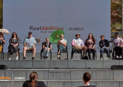 Panel discussion hosted by Nic Novice, Easterseals. Participating panelists are seated on stage. MC Danny Gomez is pointing to the audience, behind the panelits on the outdoor screen, close captioning on top reads the following; "All right. I like rolling deep. On the count of three say ReelAbilities!
