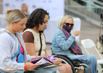 Festival attendees are sitting outdoors looking at a printed program guide. Candice Cable, Paralympian medalist is also captured in the photo looking on towards the two attendees.