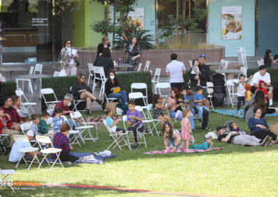 A large group of audience members of various ages. Some are seated in blankets, and others are playing outside.