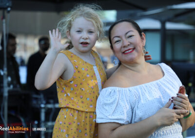 Photo of a young festival attendee waving at the camera.She is joined by a female adult both smiling and facing the camera.
