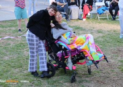 Two festival attendees one of them is leaning down, and the second person is sitting in a wheelchair. Both are smiling and looking at the camera.