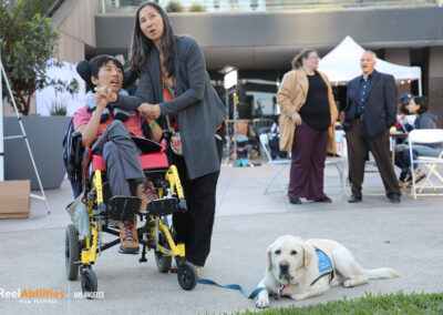 Two groups of festival attendees. The group in the foreground is a woman and a young man in a wheelchair with a service dog on a leash laying down next to them. The second group is a man and a woman engaged in conversation.