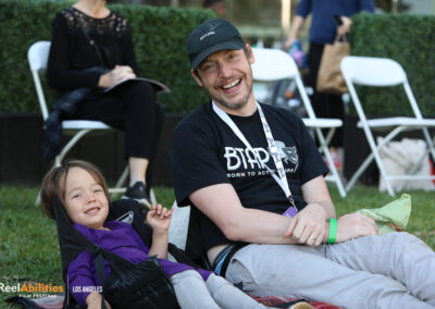 Adam Deyoe on a lawn chair with a young child seating next to him. They are both smiling, facing the camera.